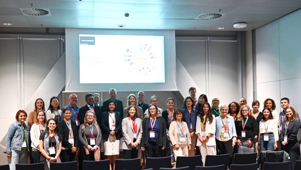 Gruppenfoto von etwa 30 Konferenzteilnehmern in einem Vortragsraum, die vor einer großen Leinwand mit einer Präsentation posieren.